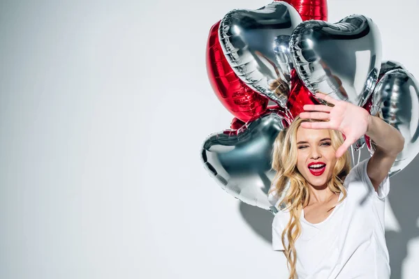 Избирательный фокус счастливой блондинки, держащей шары в форме сердца и жестикулирующей на белом — стоковое фото