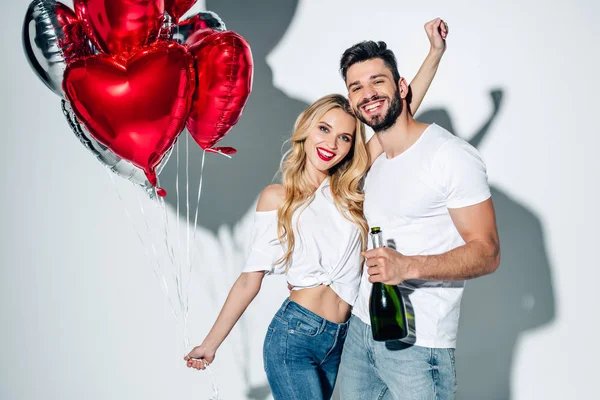 Mujer alegre sosteniendo globos cerca de hombre feliz con botella de champán en blanco - foto de stock