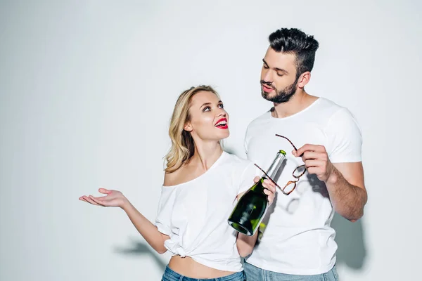 Cheerful blonde girl with bottle of champagne looking at handsome man holding glasses on white — Stock Photo