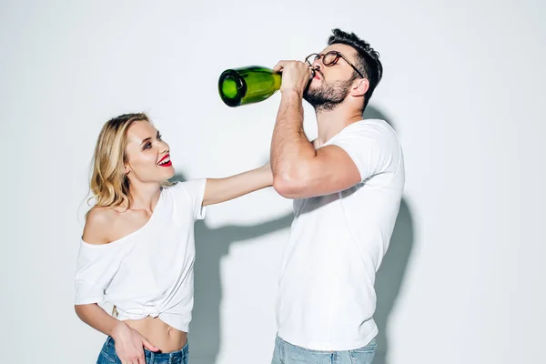 Fröhliches blondes Mädchen, das den gutaussehenden Mann in Gläsern ansieht, der Champagner aus einer Flasche auf Weiß trinkt — Stockfoto