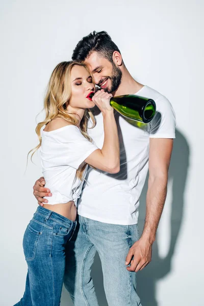 Cheerful blonde girl drinking champagne from bottle near handsome man on white — Stock Photo