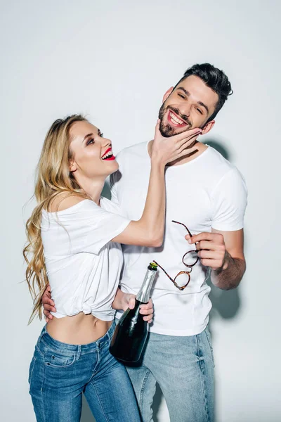 Cheerful blonde girl holding bottle and touching face of handsome man on white — Stock Photo