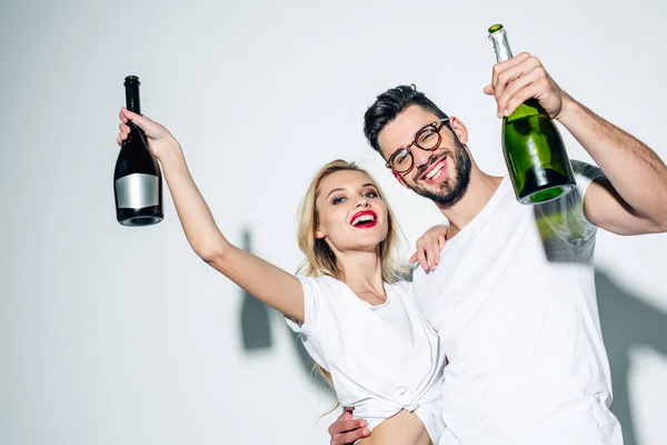 Chica rubia y hombre alegre en vasos sosteniendo botellas de champán en blanco - foto de stock