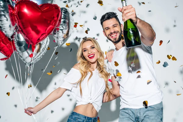 Enfoque selectivo de hombre barbudo alegre sosteniendo botella de champán cerca de la mujer con globos cerca de confeti en blanco - foto de stock