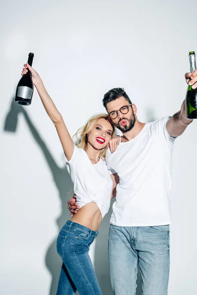 Happy blonde girl and cheerful man in glasses holding bottles of champagne on white — Stock Photo