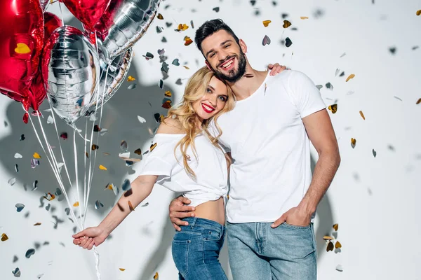 Happy bearded man hugging cheerful blonde girl with balloons near confetti on white — Stock Photo