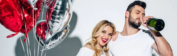 Panoramic shot of cheerful woman holding balloons near man drinking champagne from bottle on white — Stock Photo