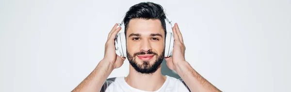 Panoramaaufnahme eines fröhlichen Mannes, der Kopfhörer berührt, während er Musik hört und auf Weiß lächelt — Stockfoto