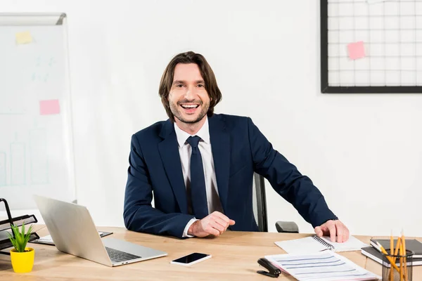 Hombre alegre en ropa formal sonriendo cerca de la computadora portátil y el teléfono inteligente en la oficina - foto de stock