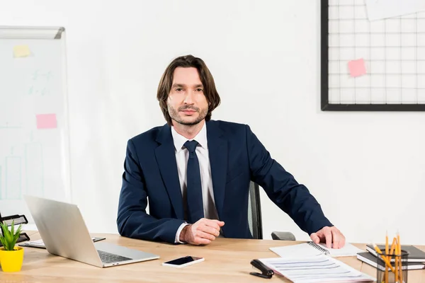 Bell'uomo in abbigliamento formale seduto vicino a laptop e smartphone in ufficio — Foto stock
