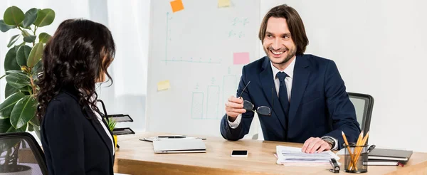 Панорамный снимок счастливого рекрутера, держащего очки и смотрящего на брюнетку в офисе — стоковое фото