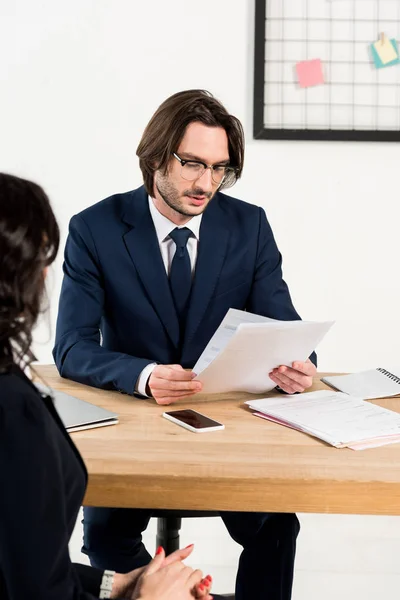 Enfoque selectivo reclutador guapo en gafas mirando documentos cerca de la mujer sentada con las manos apretadas - foto de stock