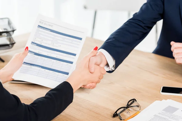 Vista recortada del reclutador sosteniendo curriculum vitae y estrechando la mano con el empleado en la oficina - foto de stock