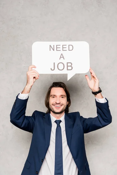 Cheerful employee holding speech bubble with need a job lettering on grey — Stock Photo