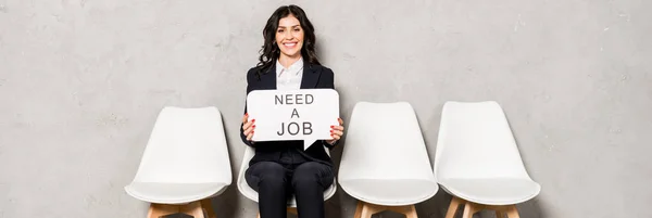 Tiro panorámico de la mujer morena feliz sosteniendo la burbuja del habla con necesidad de un trabajo de letras mientras está sentado en la silla - foto de stock