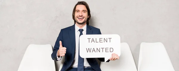 Panoramic shot of happy recruiter sitting on chair and holding speech bubble with talent wanted lettering and showing thumb up — Stock Photo