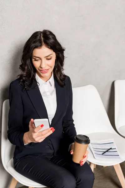 Alegre morena mujer usando smartphone mientras sostiene papel taza y sentado en silla - foto de stock