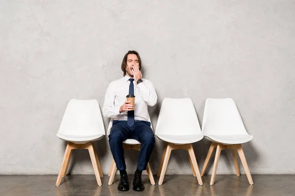 Müder schöner Mann gähnt, während er Pappbecher hält und auf Stuhl sitzt — Stockfoto