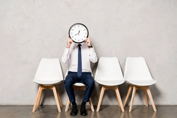 Man in formal wear covering face with clock while sitting on chair — Stock Photo