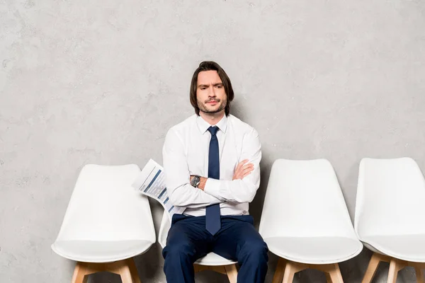 Guapo disgustado hombre sosteniendo curriculum vitae mientras sentado con los brazos cruzados - foto de stock