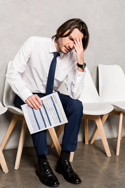 Aufgebrachter Mann in offizieller Kleidung hält Lebenslauf und berührt Gesicht, während er auf Stuhl sitzt — Stockfoto