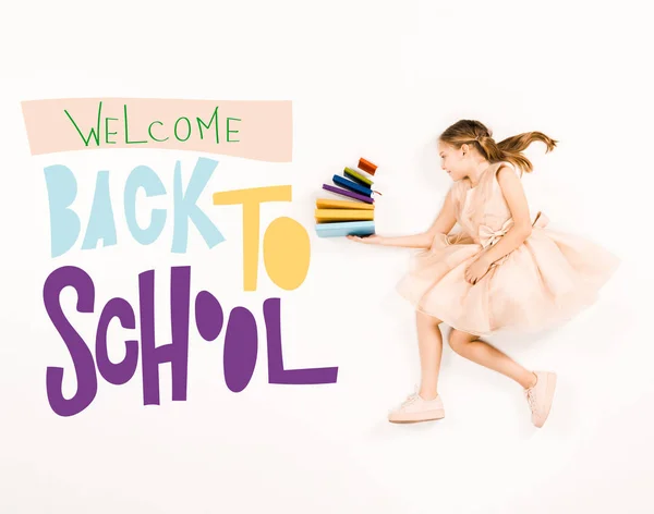 Top view of cheerful kid in pink dress holding books and flying near welcome back to school lettering on white — Stock Photo