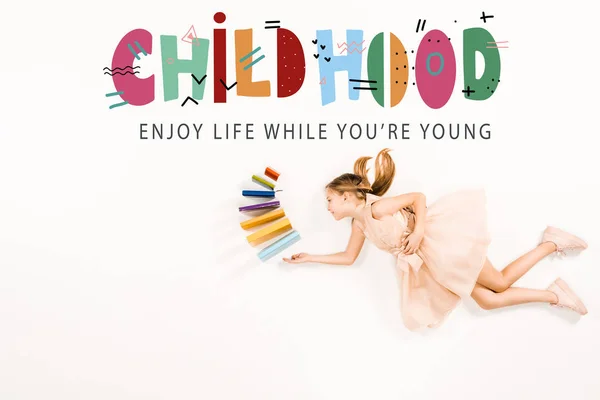 Top view of cheerful kid in pink dress holding books and flying near childhood enjoy life while you're young lettering on white — Stock Photo