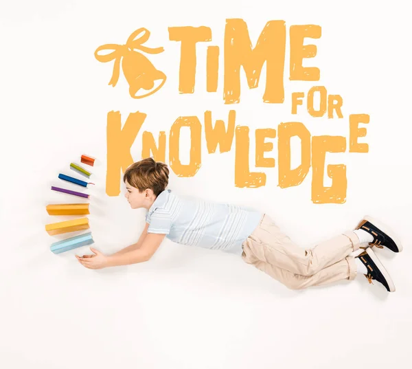 Top view of kid holding books and flying near time for knowledge lettering on white — Stock Photo