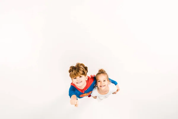 Top view of happy kid in super hero costume hugging friend and gesturing on white — Stock Photo