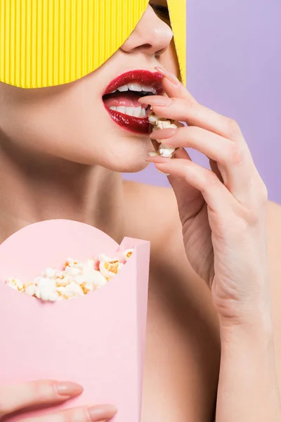 Young woman holding bucket and eating salty popcorn isolated on purple — Stock Photo