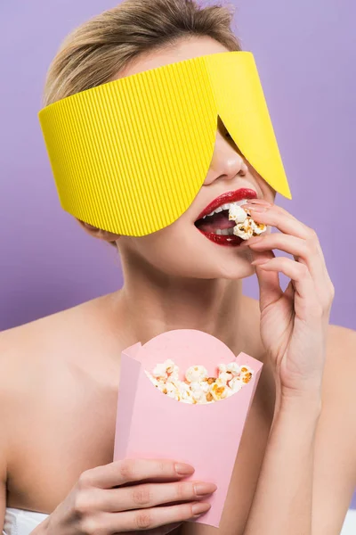 Young woman holding bucket and eating tasty popcorn isolated on purple — Stock Photo