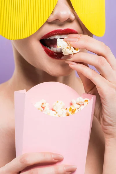 Cropped view of young woman holding bucket and eating tasty popcorn isolated on purple — Stock Photo