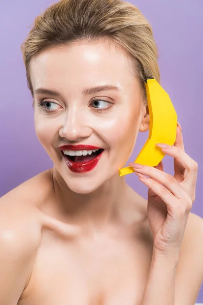 Счастливая голая молодая женщина держит желтый декоративный банан возле уха изолированы на фиолетовый — стоковое фото