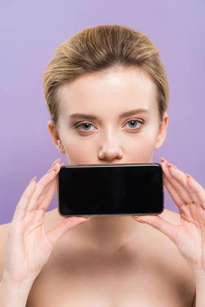 Atractiva mujer desnuda cubriendo la boca con teléfono inteligente con pantalla en blanco aislado en púrpura - foto de stock