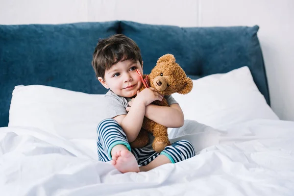 Счастливый ребенок с босиком, сидящий на кровати и обнимающий плюшевого мишку — стоковое фото