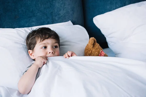 Kid lying on white pillow in bed near teddy bear at home — Stock Photo
