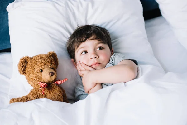 Adorable kid lying on white pillow near teddy bear at home — Stock Photo