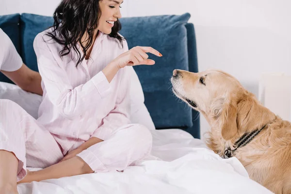 Happy woman looking at cute golden retriever while sitting on bed with man — Stock Photo