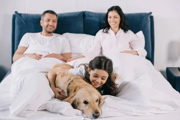 Selektiver Fokus glücklicher Kinder, die niedlichen Golden Retriever berühren, der in der Nähe der Eltern im Bett liegt — Stockfoto