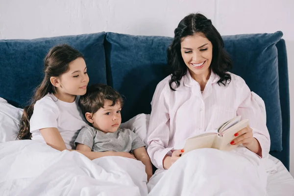 Alegre madre leyendo libro cerca de linda hija e hijo en el dormitorio - foto de stock