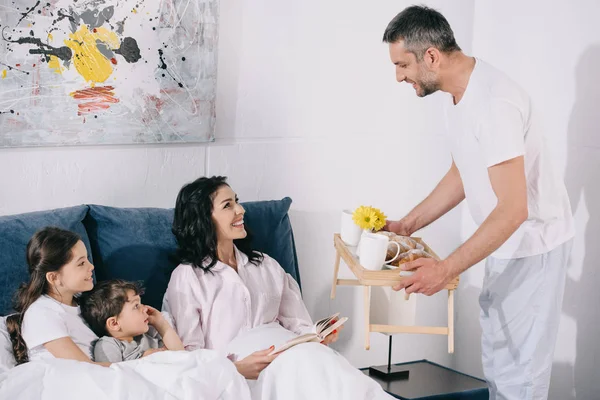 Homme heureux tenant plateau avec petit déjeuner près de la famille joyeuse à la maison — Photo de stock