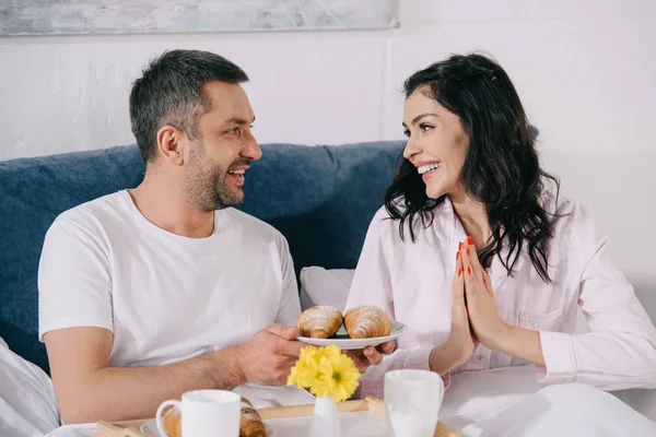 Cheerful man holding plate with croissants near happy woman in bedroom — Stock Photo