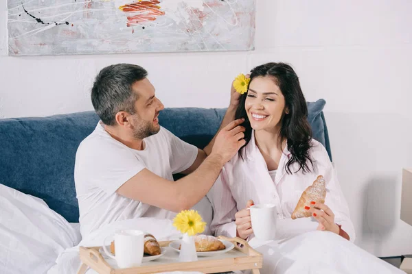 Alegre hombre sosteniendo flor cerca feliz mujer con taza y croissant en cama - foto de stock