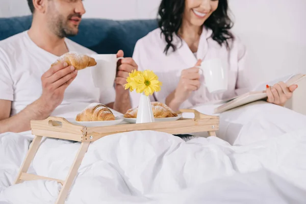 Corte vista de homem feliz segurando croissant perto de mulher lendo na cama — Fotografia de Stock