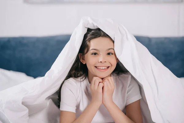 Счастливый ребенок улыбается, сидя под одеялом и глядя в камеру — стоковое фото