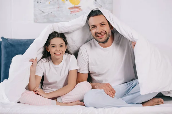 Glücklicher Vater sitzt mit süßer Tochter und hält Decke auf dem Bett — Stockfoto