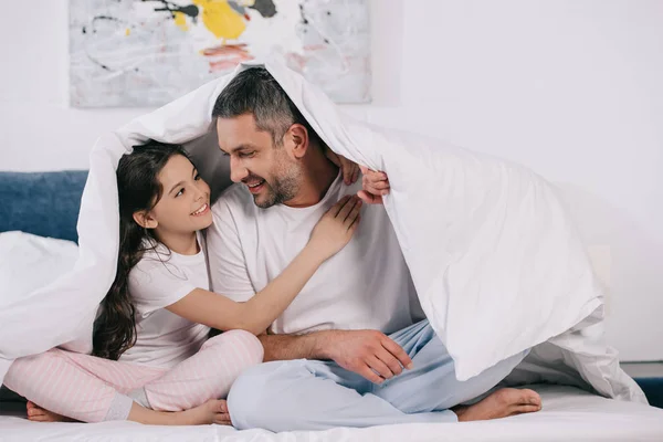 Cheerful father sitting with cute daughter and holding blanket on bed — Stock Photo