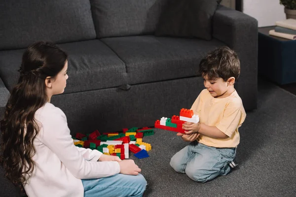 Lindo niño mirando al hermano pequeño jugando con bloques de juguete de colores en la sala de estar - foto de stock