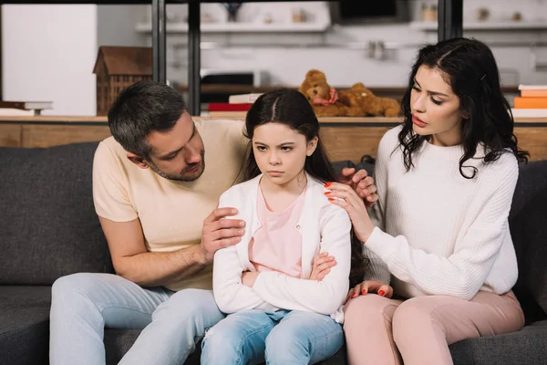 Обеспокоенные родители трогают обиженную дочь, сидящую на диване со скрещенными руками — стоковое фото