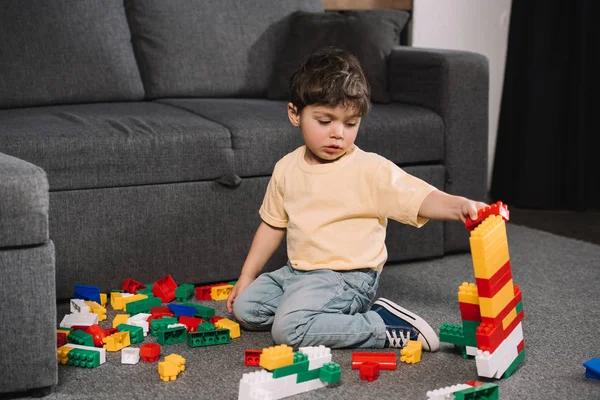Adorable niño jugando con coloridos bloques de juguete mientras está sentado en el suelo en la sala de estar - foto de stock
